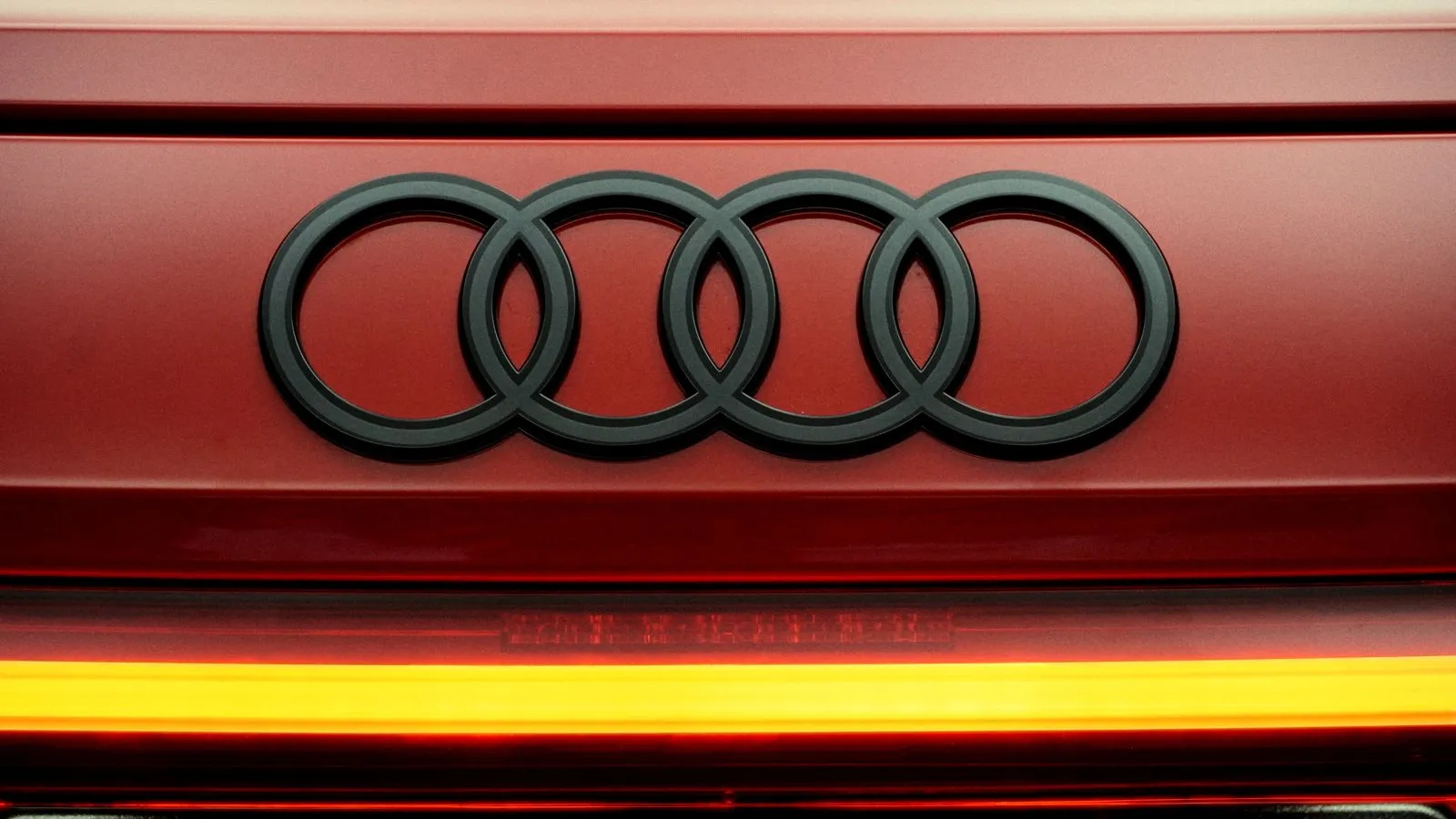 Close-up of the black four-ring Audi badge on the red tailgate of a car, with a lit amber light strip below, no people