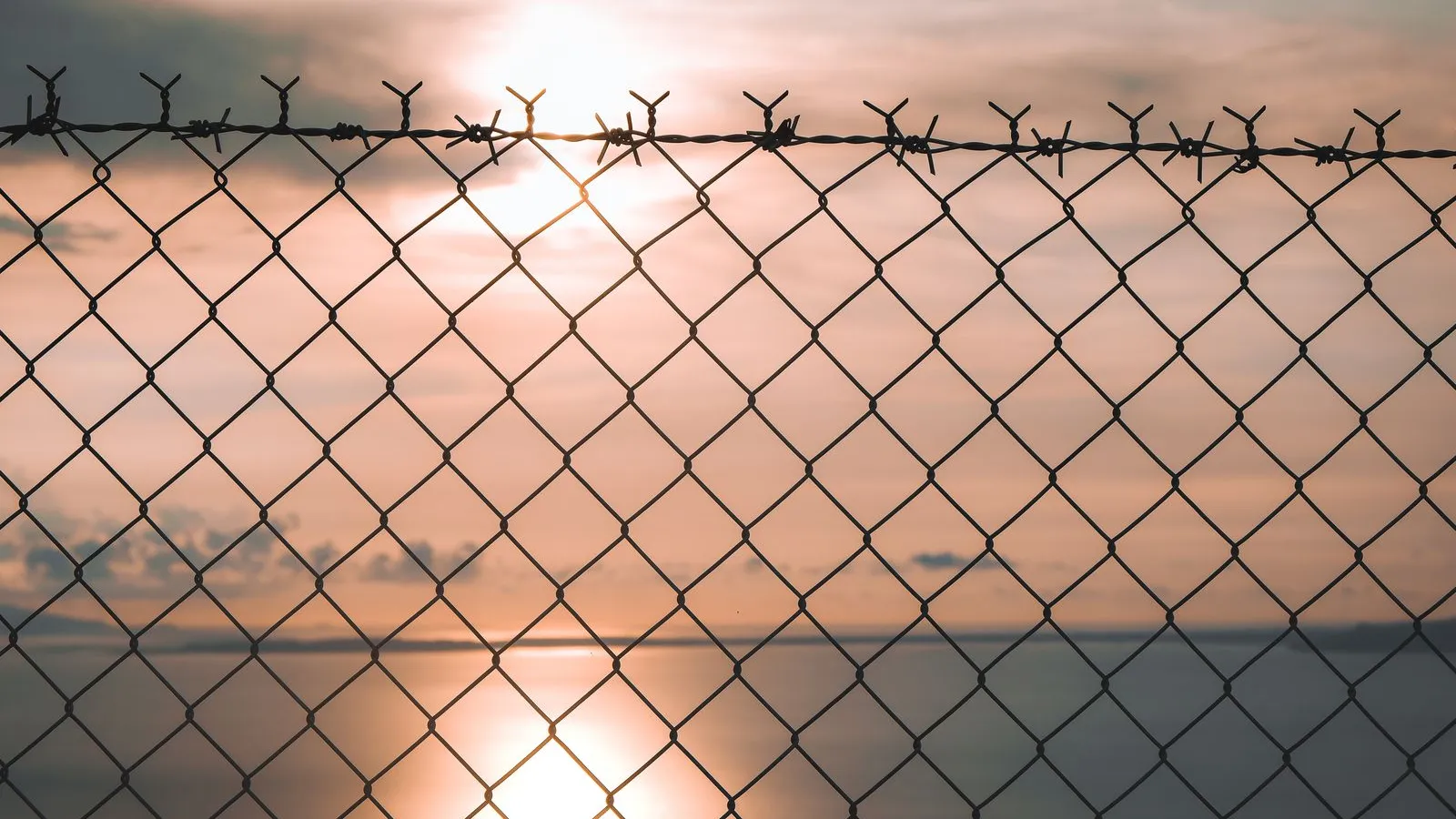 A chain-link fence topped with barbed wire against a sunset over the sea — file stock photo, not the Ceuta border fence, no people