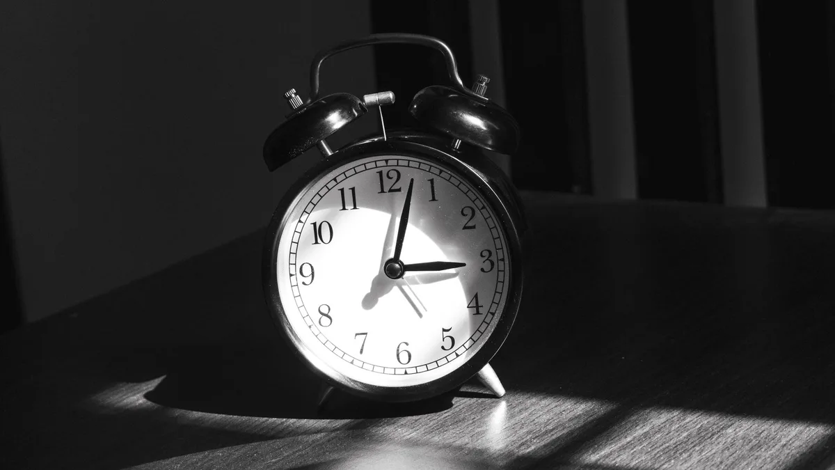 A black vintage twin-bell alarm clock showing just after three o'clock, in black-and-white light and shadow on a wooden table — file stock photo, no people