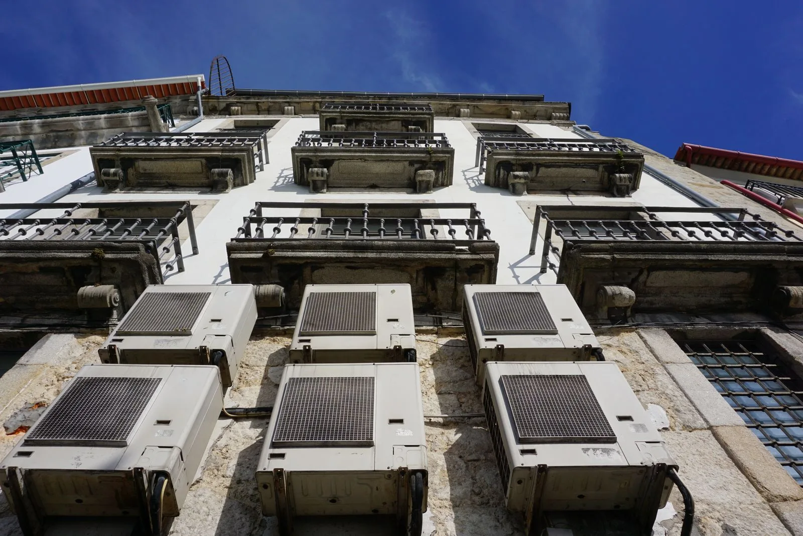 Air-conditioning units mounted on the stone facade of an old apartment building in Porto, seen from below against a blue sky, no people