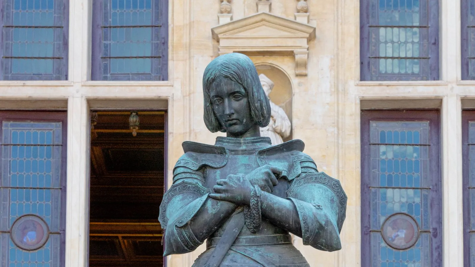 Bronze statue of Joan of Arc in armour, hands crossed on her chest, in front of the stone facade and stained-glass windows of the Hôtel Groslot in Orléans, no people