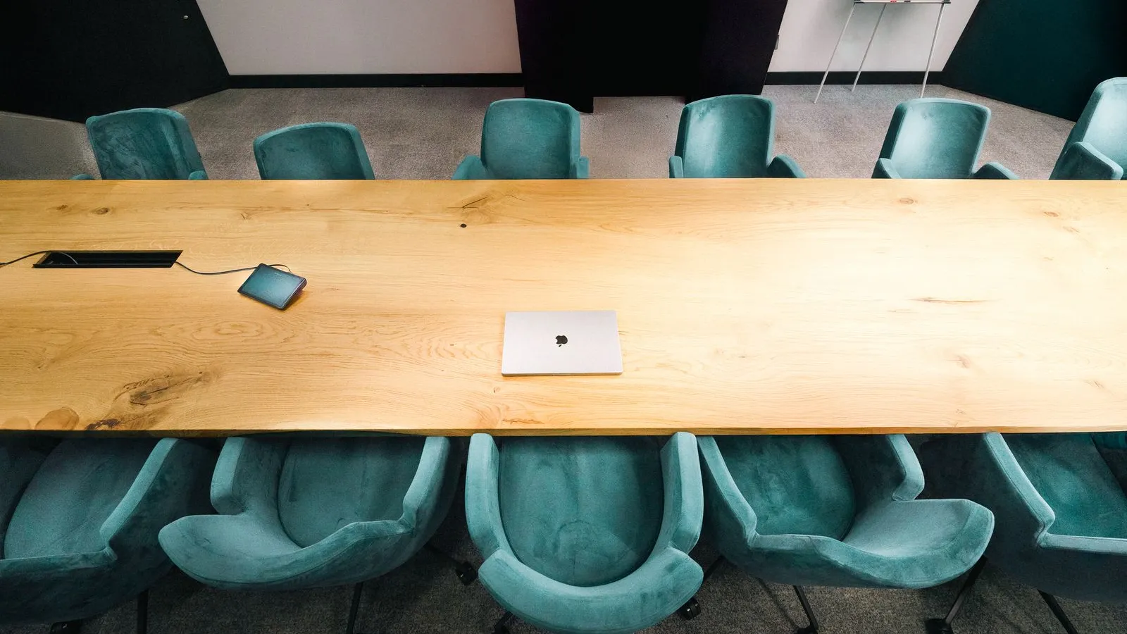 A long, empty light-oak conference table with teal upholstered chairs on both sides and a closed laptop in the middle, no people