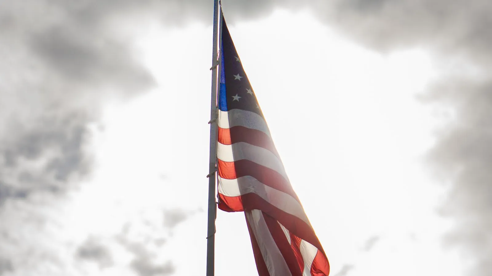 The flag of the United States on a flagpole, backlit against heavy grey clouds — file stock photo, not taken at a US base in Europe, no people