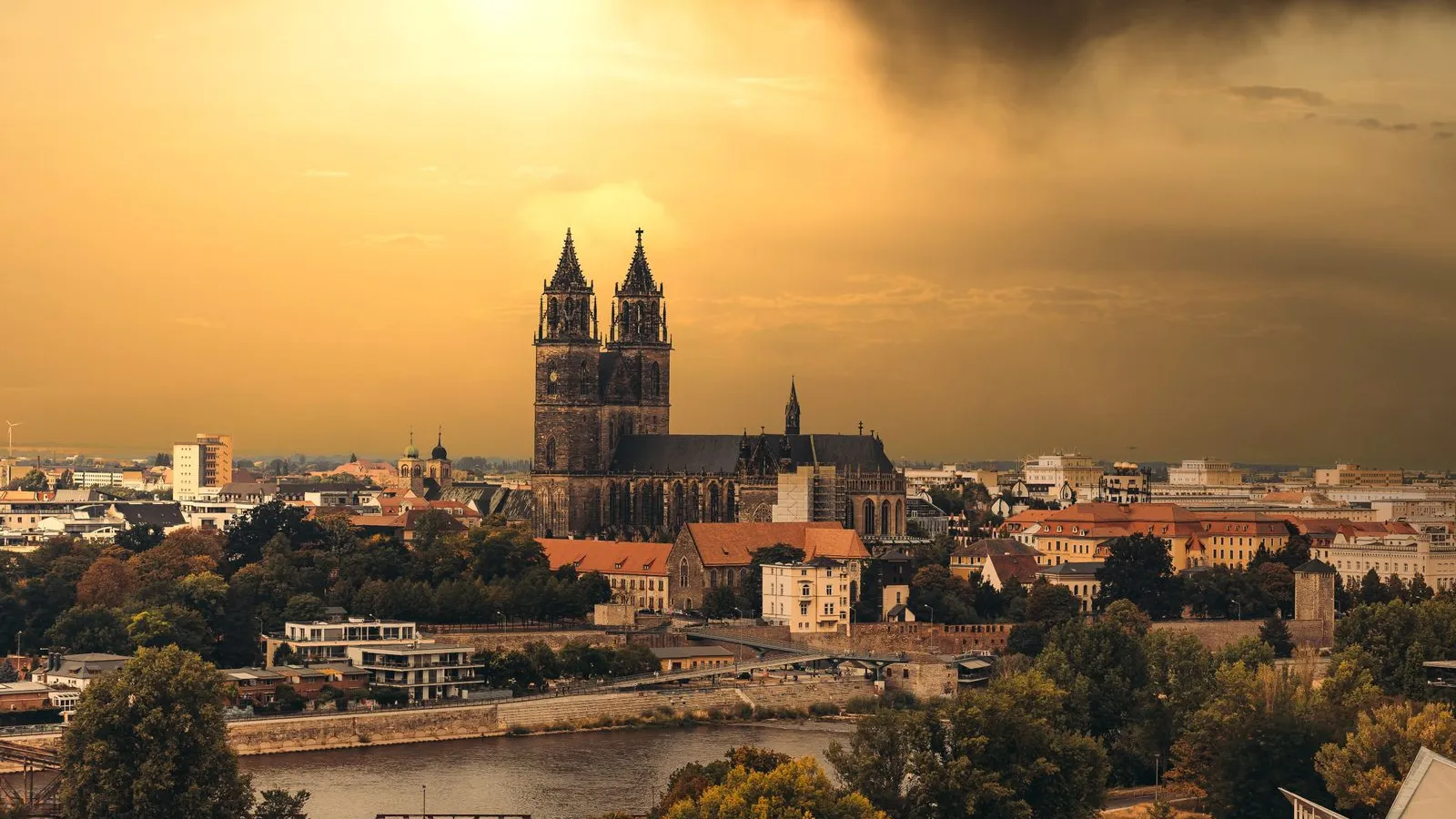 Magdeburg Cathedral and the city skyline above the Elbe under a dark storm sky at golden hour; the Saxony-Anhalt state parliament sits on the cathedral square — file stock photo, no identifiable people