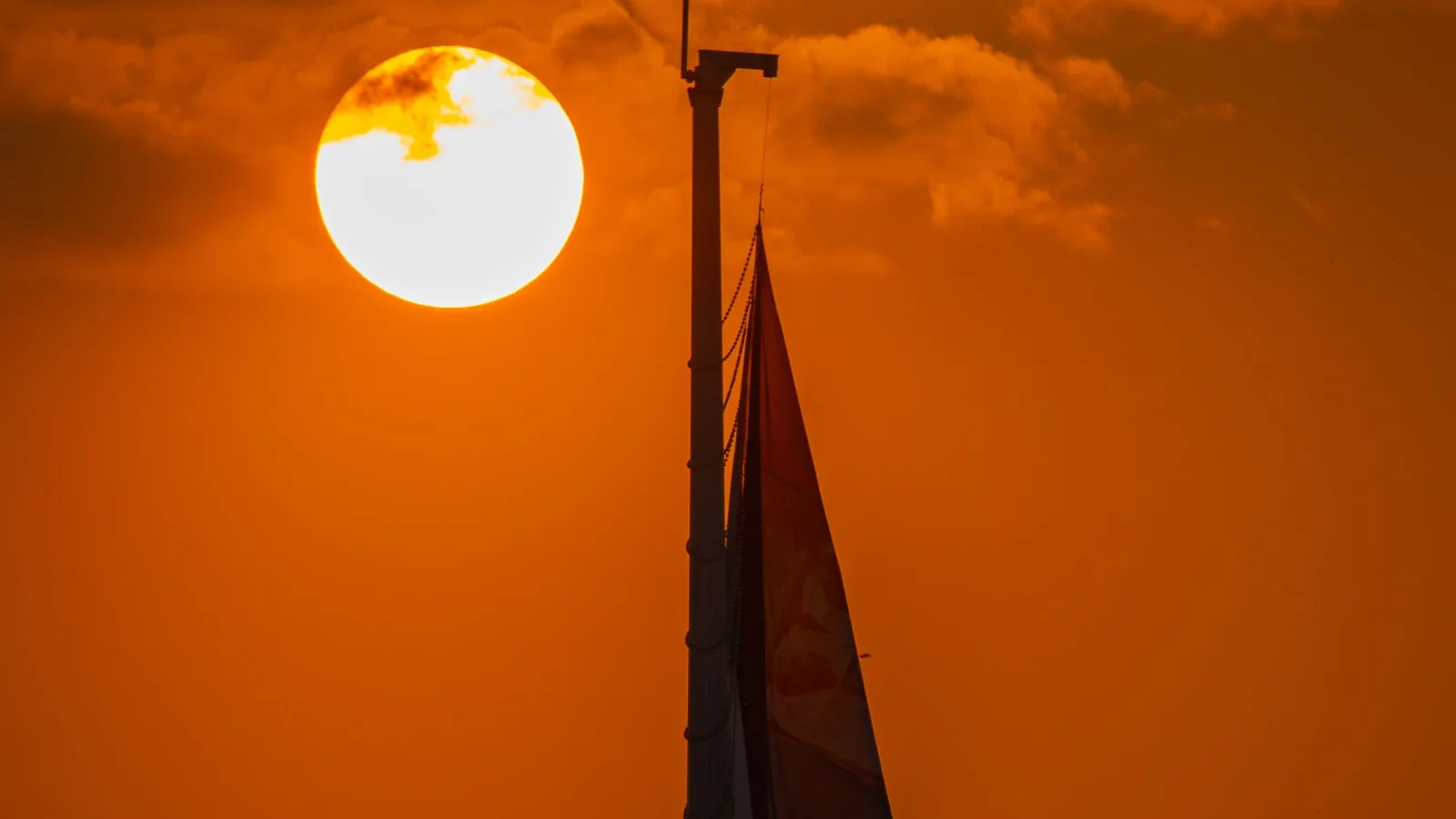Silhouette of a flagpole and a furled Serbian flag against a large setting sun and an orange sky over Belgrade, no people