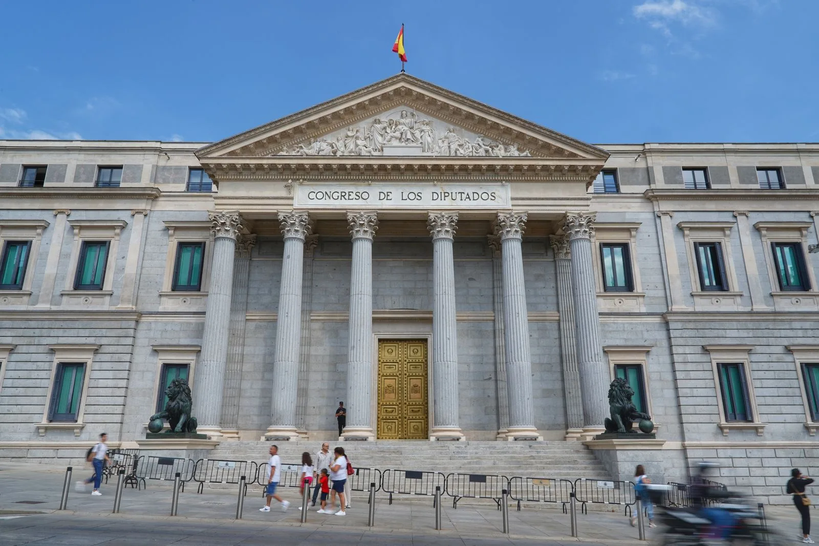 The neoclassical façade of the Congreso de los Diputados in Madrid with its bronze lions and the Spanish flag on the roof, under a blue sky — file stock photo, passers-by not identifiable