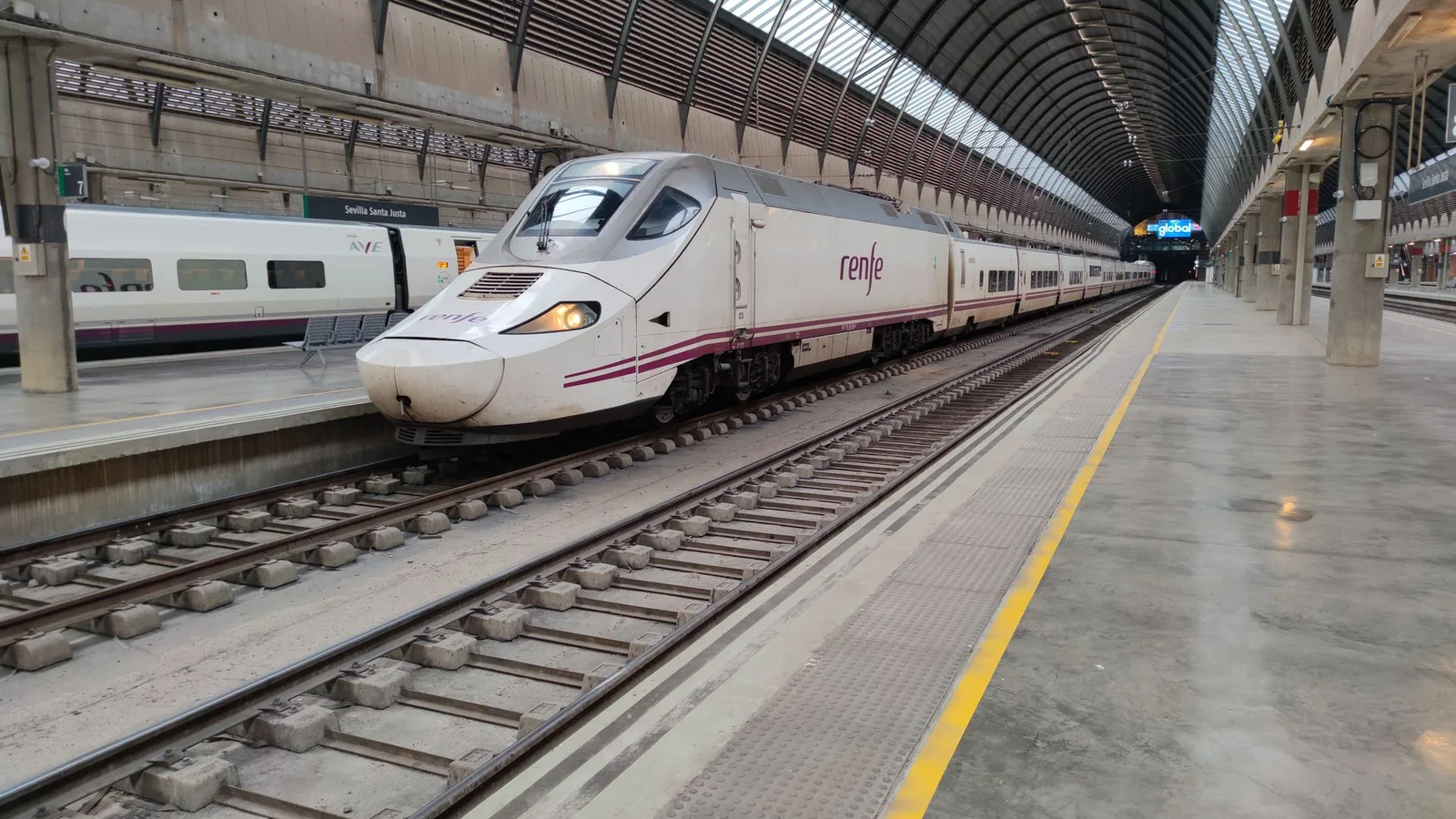 A white Renfe high-speed train with a purple stripe standing at a platform under the arched roof of Seville Santa Justa station, no people; file stock photo, not one of the trains involved in the crash