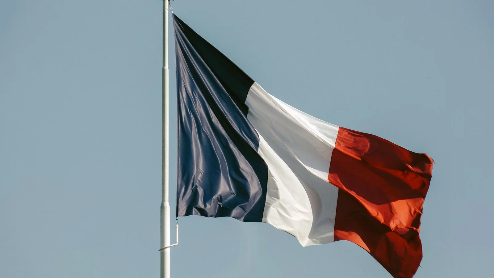 The French tricolour flag flying from a white flagpole against a clear pale blue sky, no people