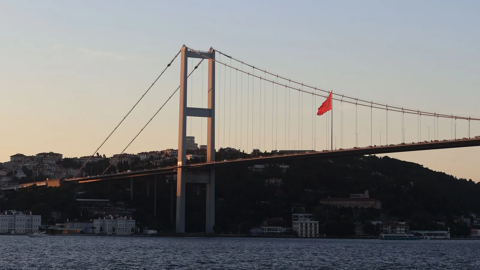 The Bosphorus Bridge in Istanbul at sunset with a Turkish flag flying on the shore — file stock photo published on Pexels in June 2026, no people