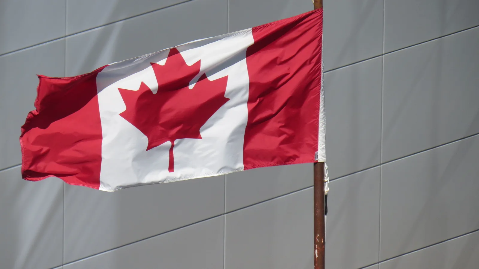 A red-and-white Canadian maple leaf flag flying from a flagpole in front of a grey modern building facade, no people; file stock photo, not taken at the Montreal summit